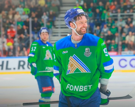 Хоккеисты в зелёной форме находятся на льду во время матча. Один из игроков улыбается, в руках у него клюшка. На заднем плане видна трибуна с болельщиками